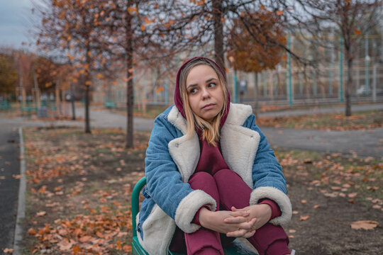A woman sitting pensively on a bench in a park surrounded by trees during autumn