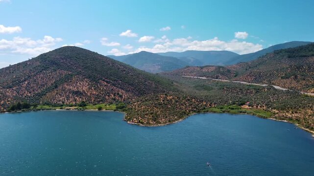 An aerial shot captures the tranquil blue waters of Bafa Lake, nestled amidst the lush, tree covered hills and mountains of Turkey. The clear sky with scattered clouds enhances the natural beauty of t