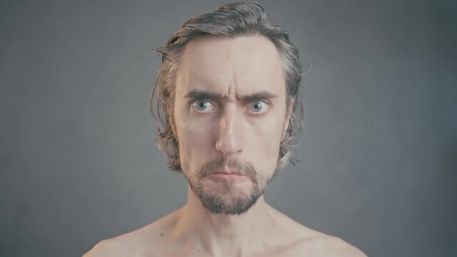 Funny man covers his mouth with hands, showcasing intense facial expressions in a minimalistic setting and serious emotions against a neutral gray background.