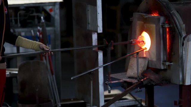 Close-up glassblowing process tube tool with molten glass shaped inside refractory furnace in industrial craft workshop. Handmade blowing glassware design production manufacture Europe
