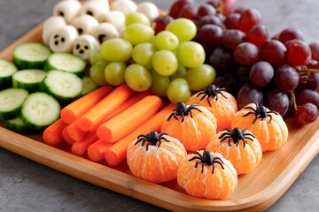 Vibrant fruit and vegetable platter with peeled oranges, grapes, cucumbers, carrots, and Halloween-style spider-topped mandarins on a wooden board.