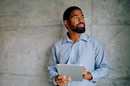 Black businessman holding a digital tablet, looking away with a thoughtful expression, standing against a concrete wall