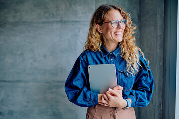 Businesswoman standing by window in modern office, holding a digital tablet, looking away and smiling © we.bond.creations