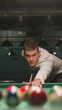 Close-up of billiard player in white shirt and grey trousers gripping cue stick with intense focus. Green pool table with scattered billiard balls. Overhead lights cast dramatic shadows
