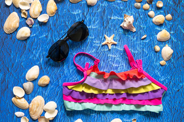 A collection of travel items including suitcase, sunglasses, swim suit, sunscreen and shells on turquoise navy blue background