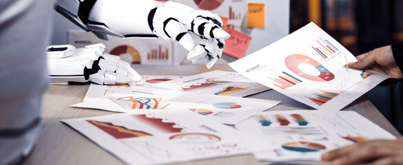 A robotic arm examines colorful business charts and graphs in a modern office setting, showcasing the intersection of technology and data analysis for enhanced decision-making.Noogenesis