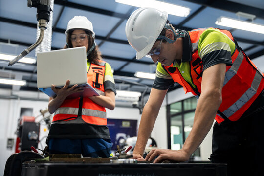 Workers in high-visibility vests coordinating factory tasks with laptop and robot machinery automation, engineering innovation, safety, and quality control in modern industry