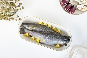 Two fresh fishes lie in a white plate with lemon wedges; salt and zest coat their skin. Next to them sit fresh eggplants and dried herbs on a white surface
