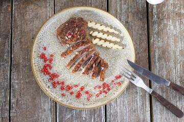 A perfectly cooked duck breast, sliced and adorned with pomegranate seeds, is presented with grilled baby corn on a speckled plate, ready to be savored with a knife and fork © Damian