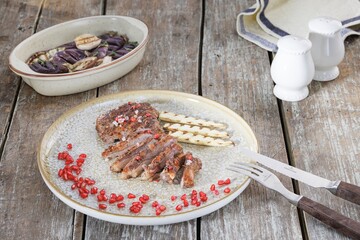 A plate of sliced meat with grilled baby corn and pomegranate seeds is presented. Next to the plate sits a dish of eggplant and salt and pepper shakers on a weathered wooden table