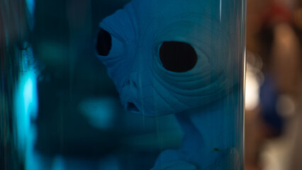 A detailed alien head with large black eyes is displayed inside a glass case, bathed in eerie blue...