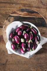 A collection of small, purple eggplants, some with their green stems, rests in a white bowl lined with linen, sitting on a wooden surface and a round cutting board