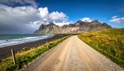 A scenic coastal landscape featuring a dark sandy beach, winding dirt road, and towering mountains partially veiled in clouds. Vivid blue sky overhead
