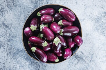 Many small, shiny, purple eggplants are piled into a black bowl. The bowl sits on a textured grey and white surface, ready to be used in a delicious recipe