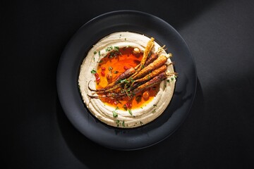A plate presents a visually appealing dish of creamy hummus, drizzled with vibrant orange sauce and adorned with roasted carrots, herbs, and tiny red pomegranate seeds