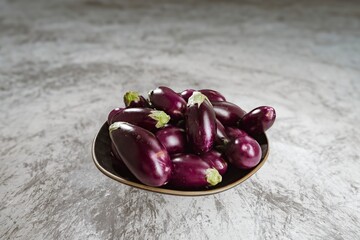 A collection of shiny, dark purple eggplants, each with a small green stem, fills a simple dark brown bowl. The bowl sits on a mottled grey and white background