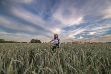 Fototapeta premium Young woman with long hair stands confidently in a golden wheat field at sunset, showcasing a serene rural landscape with vibrant sky colors and natural beauty