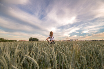 Fototapeta premium Young woman with long hair stands confidently in a golden wheat field at sunset, showcasing a serene rural landscape with vibrant sky colors and natural beauty