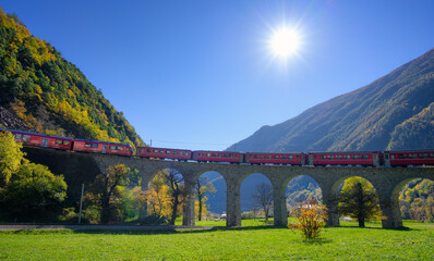 Bernina Express red train on Brusio spiral viaduct in Swiss alpine mountains sunny autumn day. Kreisviadukt, Switzerland. Train on the bridge in Alps. Railroad, green grass , colorful trees in fall