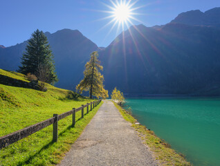 Scenic view of a walking path along turquoise lake in Switzerland with bright sunlight over alpine mountain peaks in fall. Green grass and autumn trees create a peaceful colorful landscape. Nature
