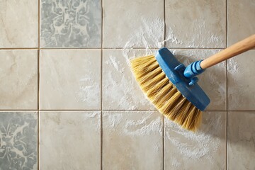 A close-up, overhead view of a broom sweeping white powder and dust from light-colored ceramic tiles.