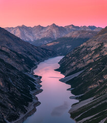 Aerial view of a mountain valley with a lake at sunset in the Italian Alps. Pink evening sky over rocky peaks and calm water. Top view of rocks, hills, canyon, reflection in water and pink sky at dusk