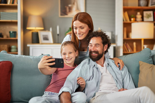 A joyful family of three captures a fun moment using a smartphone while relaxing on a comfortable couch in their inviting living room filled with warm sunlight.