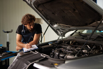 Mechanic assesses a vehicle's engine while making notes on a clipboard in a busy car repair shop. Tools and equipment are visible in the background.