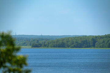 Calm lake with forested shoreline and distant hills under blue sky