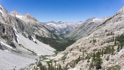 Aerial view of a mountain valley with snow capped peaks and a dense forest under a clear blue sky