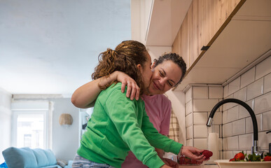 Affectionate daughter kissing her mother on the cheek while washing strawberries together in the kitchen at home