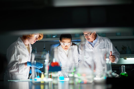 Two students in white coats and protective glasses conducting chemical experiment in university lab under the control of their professor