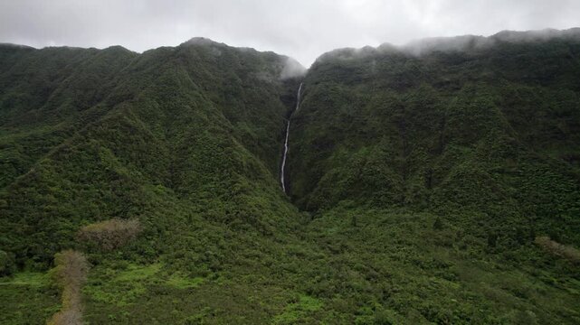 Cinematic drone orbit over Bibron waterfall on Reunion Island. A tall, narrow cascade descends dramatic cliffs covered in lush tropical vegetation, with drifting clouds enhancing the humid jungle feel