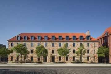Historic stone building showcases elegant architecture under clear blue sky with trees lining the walkway, situated in an urban area during daytime