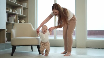 Baby development. First steps. Little infant girl making first steps at home. Happy mother supporting baby first steps. Mom helping toddler child walking in living room. Happy family at home