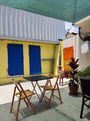 A vibrant and eclectic outdoor patio scene featuring a small black metal bistro table with two light wooden folding chairs, set against a bold, colorful architectural backdrop 