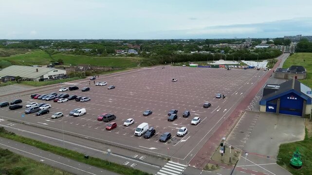 Aerial view of large parking lot with scattered cars, blue entrance building, crosswalk, and nearby residential area highlighting infrastructure and spatial layout.