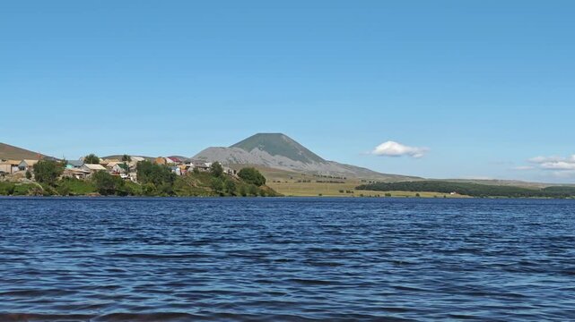 Serene lake water ripples gently with Mount Aragats and a village nestled on the shore under a clear blue sky