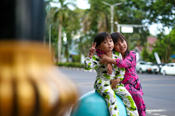 Two little girls in colorful clothes sitting on a blue round object outdoors, hugging and smiling,...