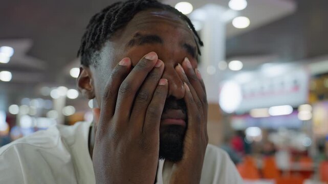 student seated inside busy restaurant shows sign of exhaustion with eyes rolled upward and lips pressed in frustration while holding hands together, surrounded by people in blur background