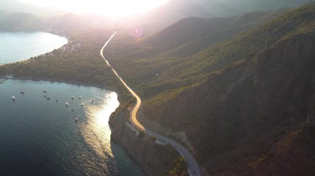 An aerial view captures the scenic coastal road winding along the Dat&ccedil;a Peninsula, leading towards the historic ruins of Knidos Ancient City in Turkey. The golden hour light illuminates the winding pa