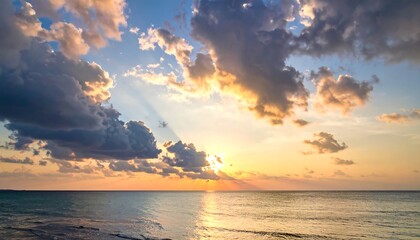A tranquil seascape captures a golden sunset. Clouds billow across the sky, sunlight piercing through. Calm ocean waters reflect the warm glow