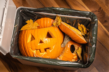 Discarded Halloween pumpkins with carved faces and autumn leaves are placed in a trash bin, showcasing seasonal waste and the end of festive celebrations