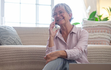 Happy senior woman sitting at home talking on smartphone. Elderly woman smiling and enjoying relax with technology, online communication, and digital lifestyle in a cozy living room.