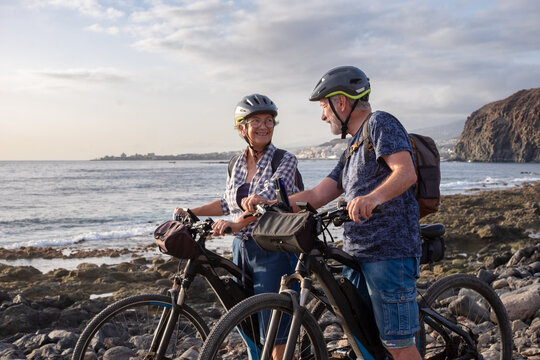 Senior active couple riding on the pebble beach with electric bicycles at sunset. Authentic elderly retired life and sustainable mobility concept