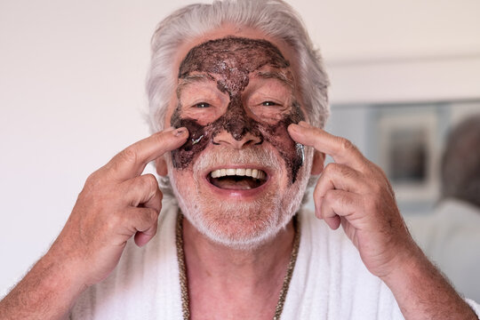 Skin care. Cheerful bearded senior man in bathrobe applying black scrub on his face looking at camera smiling - Powered by Adobe