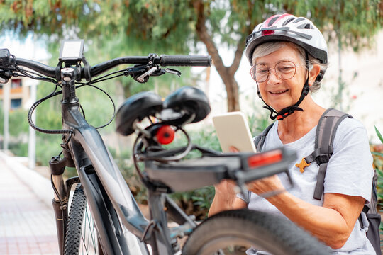 Smiling senior caucasian cyclist woman with helmet in leisure activity in urban park riding with electric bike resting using smartphone