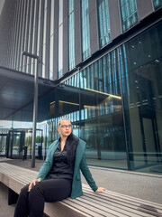 Pretty young caucasian woman in stylish green suit sitting near modern office building, summer day