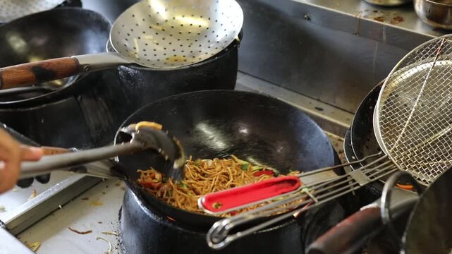 Chef stir frying Asian noodles with vegetables in hot wok at restaurant kitchen, with steam and flame.