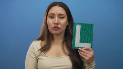 Young hispanic woman holds reflective green learner plate beside her face in a blue studio; determination.